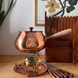 Peut inclure: Un service à fondue en cuivre vintage, comprenant une marmite avec une poignée et un couvercle en bois, un support en laiton, un brûleur et un plateau. Comprend des fourchettes à fondue avec des embouts colorés.