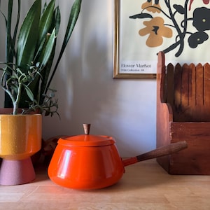 May include: A vibrant orange enamel saucepan with a wooden handle and lid. The pan sits on a wooden surface, with a plant in a yellow and pink pot to the left. A framed floral print is in the background.