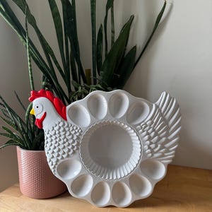 Peut inclure: Un présentoir à œufs en céramique blanche en forme de poule. Il comporte des emplacements pour une douzaine d'œufs et un bol central. La poule a une crête et un fanon rouges, et un bec jaune. Le présentoir est sur une surface en bois.