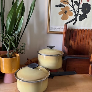 Peut inclure: Deux casseroles en émail jaune avec des poignées et des couvercles noirs. Les casseroles ont des bords argentés et des boutons noirs. Un coupe-pain en bois et une affiche florale encadrée sont en arrière-plan.