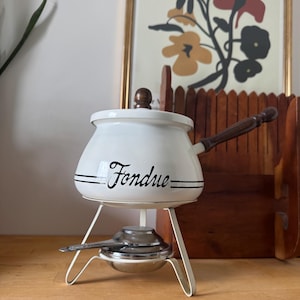 May include: White enamel fondue pot with a wooden handle and lid. The pot has the word "Fondue" in black script with two black lines. It sits on a white metal stand with a burner and a small spoon. A vintage kitchenware item.
