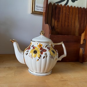 May include: A white ceramic teapot with a floral sunflower design in yellow, orange, and brown. The teapot has a gold-coloured handle, spout, and trim. The lid is topped with a gold finial. The teapot is sitting on a wooden surface.