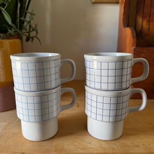 May include: Four white ceramic mugs with a blue grid pattern. Each mug has a rounded handle and a stacked design. The mugs are arranged in two stacks of two on a wooden surface.
