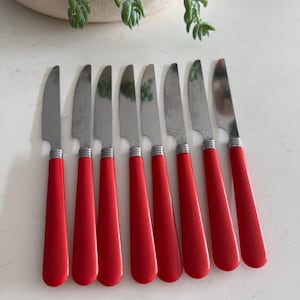 Peut inclure: Un ensemble de huit couteaux à beurre en acier inoxydable, avec des manches rouges vifs. Les couteaux sont disposés en rangée, les lames orientées dans la même direction. Les manches sont rouge brillant, et les lames sont argentées.