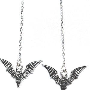 May include: A pair of silver-colored bat-shaped earrings. Each earring features a detailed bat pendant hanging from a delicate chain. The bats have outstretched wings and intricate detailing.