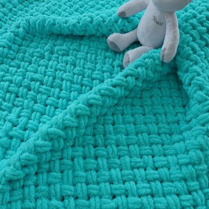 May include: A bright turquoise chunky knit blanket with a thick, woven texture. A light gray plush teddy bear with embroidered details on its chest sits on top of the folded corner of the soft, yarn-based textile fabric.