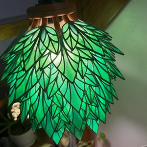 May include: A green stained glass pendant lamp with a leaf design. The lamp shade is made up of many green glass leaf shapes, outlined in black. The lamp hangs from a wooden fixture. The light is on, illuminating the leaves.