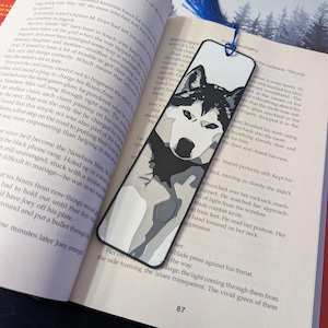以下が含まれることがあります： ハスキー犬のイラストが描かれた白黒のブックマーク。青いタッセルが付いており、「HELLBENT」というタイトルの本に挟まれています。