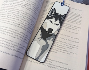 Puede incluir: Marcapáginas en blanco y negro con una ilustración de un perro husky. El marcapáginas tiene una borla azul y está colocado en un libro abierto con el título "HELLBENT".