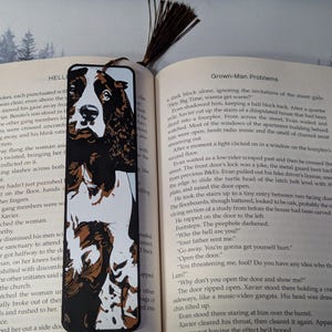 May include: A black, white, and brown illustrated bookmark featuring a dog's face. The bookmark is rectangular with a tassel and is placed in an open book with text.