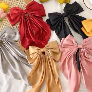 Könnte beinhalten: Eine Sammlung großer Haarschleifen aus Stoff in verschiedenen Unifarben wie Rot, Schwarz, Gelb, Silber, Gold und Rosa. Jede Schleife hat eine strukturierte Knotenmitte mit langen, fließenden, gefalteten Bändern, die nach unten hängen.