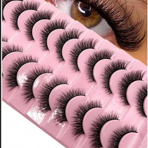 Könnte beinhalten: Ein Set schwarzer falscher Wimpern, angeordnet auf einem rosa Tablett. Die Wimpern sind lang und voll, mit einem natürlich aussehenden Schwung. Das Bild zeigt auch eine Nahaufnahme eines braunen Auges mit langen, dunklen Wimpern.