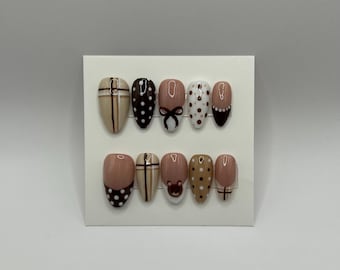 May include: A set of ten almond-shaped false nails. The nails feature various designs, including polka dots, bows, and stripes, in shades of brown, beige, and white. Some nails have a matte finish, while others are glossy. The nails are arranged on a white card.
