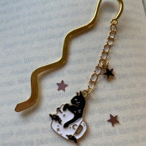 May include: Gold-colored metal bookmark with a wavy top. A chain with a black cat in a white teacup charm and two black star charms hangs from the bottom. The cat charm has black and white enamel details. Decorative stars are scattered around the bookmark.