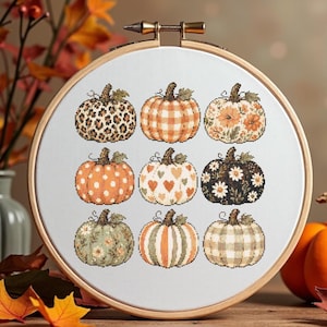 Peut inclure: Un tambour à broder circulaire en bois contenant un tissu blanc avec un motif au point de croix représentant neuf citrouilles. Les citrouilles présentent divers motifs, notamment des imprimés léopard, des carreaux orange, des fleurs et des cœurs.