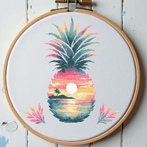 May include: A circular wooden embroidery hoop containing white fabric with a cross-stitch design of a pineapple. The pineapple features a tropical sunset scene inside its body, with a palm tree, a white sun, and water, flanked by two small pink plants.