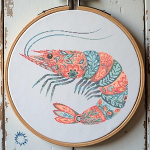May include: A circular wooden embroidery hoop containing white fabric with a cross-stitch design of a shrimp. The shrimp features a detailed floral and leaf pattern in shades of coral, peach, teal, and dark blue, with long thin antennae.