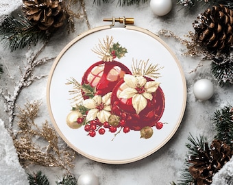 May include: A cross-stitch embroidery hoop featuring a Christmas ornament design. The design includes two red ornaments, white poinsettias, and red berries. The hoop is surrounded by pine needles, pine cones, and white ornaments.