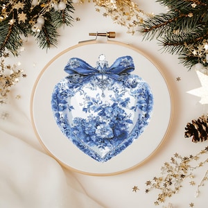 以下が含まれることがあります： 木製の刺繍枠に入ったハート型のクロスステッチデザイン。デザインは青と白の花柄で、青いリボンが飾られています。背景には、松の枝や金の星のアクセントなど、クリスマスの装飾が含まれています。