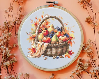 Puede incluir: Un bastidor de bordado con una imagen detallada de una cesta llena de frutas de otoño. La cesta contiene manzanas, peras y uvas, rodeadas de follaje otoñal. La paleta de colores incluye naranjas, rojos y marrones cálidos.