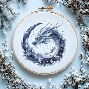 Könnte beinhalten: Kreuzstich-Stickrahmen mit einem blau-weißen Drachendesign. Der Drache ist von Schneeflocken und einer winterlichen Landschaft umgeben. Die Stickerei befindet sich in einem Holzrahmen mit einer Messingschraube.