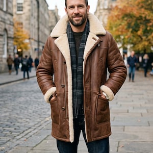May include: A brown leather coat with a thick cream-coloured shearling lining, a wide notched collar, and long sleeves with shearling cuffs. The coat features a button-front closure, two front pockets, and is worn over a dark green sweater and scarf.