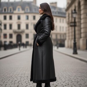 May include: A long, black leather coat with a shearling lining and a belted waist. The coat is worn by a person standing on a cobblestone street in front of a European-style building.