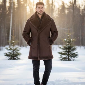 May include: A brown shearling coat with a wide collar and cuffs. The coat has a button-up front and is worn over a dark shirt and black pants. The person is standing in a snowy forest with evergreen trees in the background.