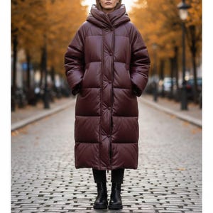 May include: A long, burgundy puffer coat with a large hood and button closure. The coat is worn over black trousers and boots. The coat is a solid colour and appears to be made of a leather-like material. The background features a tree-lined street with autumn foliage.