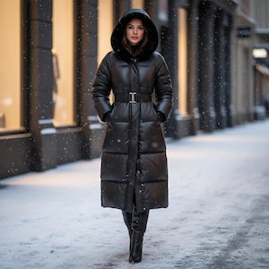 May include: A long, black puffer coat with a hood and belt, worn in a snowy outdoor setting. The coat is knee-length and has a belt at the waist. The model is wearing black boots. The background shows buildings and falling snow.