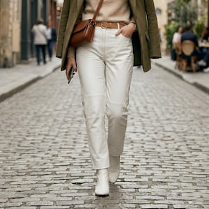 May include: A person wearing a tan sweater, white pants, and white boots walks down a cobblestone street. They are wearing a brown leather belt and carrying a brown leather purse. A green jacket is draped over their shoulders.