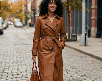 May include: A woman wearing a brown leather trench coat with a matching tote bag. The coat has a belted waist, button details, and a classic trench coat design. The woman is walking on a cobblestone street.