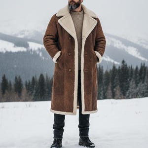 May include: A long brown coat with a cream-colored shearling lining and trim. The coat has a double-breasted closure and two pockets. The person in the image is wearing black jeans and black boots. The background is a snowy landscape with evergreen trees.