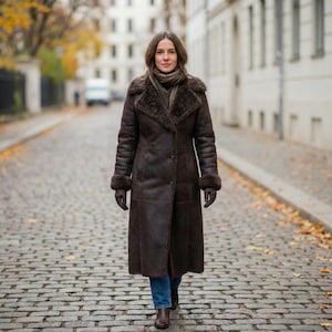 May include: A long, dark brown shearling coat with a fur collar and cuffs. The coat is buttoned up and worn with gloves and boots. The person is walking on a cobbled street.