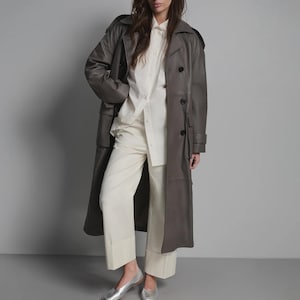 May include: A long, gray leather trench coat with a double-breasted design. The coat is worn over a white button-down shirt and cream-colored pants. Silver flats complete the outfit, creating a sophisticated, minimalist look.