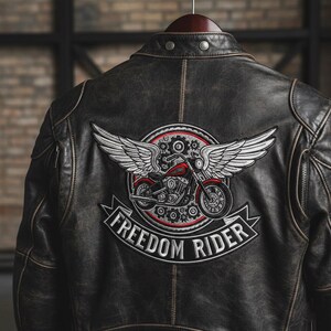 Peut inclure: Veste en cuir noir vieilli avec un grand écusson brodé dans le dos. L'écusson représente une moto rouge ailée, entourée d'un cercle rouge, avec l'inscription "FREEDOM RIDER" en blanc.