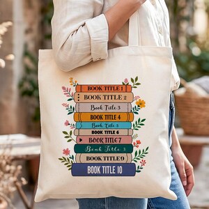 Könnte beinhalten: Eine cremefarbene Stofftasche aus Segeltuch mit einem Grafikdruck eines Stapels aus zehn bunten Büchern. Jeder Buchrücken ist mit dem Text BOOK TITLE 1 bis BOOK TITLE 10 beschriftet und von kleinen, bunten Blumenillustrationen umgeben.