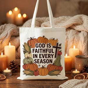 May include: A cream-colored canvas tote bag featuring a graphic print of pumpkins, corn, apples, and a cross. The text "GOD IS FAITHFUL IN EVERY SEASON" is printed in black in the center. The bag has two shoulder straps and a rectangular shape.