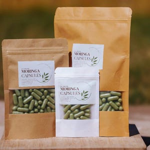 May include: Three packets of Moringa capsules in different sizes. The capsules are green and visible through clear windows in the packaging. The packaging is a mix of brown paper and white plastic, with labels that read "Moringa Capsules".