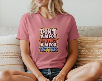 May include: A dusty rose t-shirt with the text "Don't Aim for Perfect, Aim for Better" in a colorful, retro font. The shirt is a crew neck and is being worn.