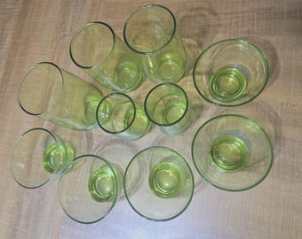 May include: A collection of clear glass drinkware with a subtle green tint. The assortment includes various sizes of glasses and bowls, arranged on a light-colored wooden surface. The glassware is designed with a simple, elegant aesthetic.