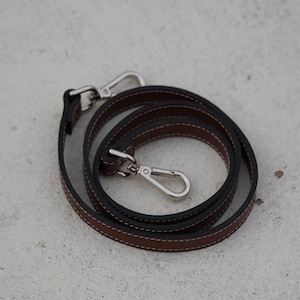 May include: A coiled brown leather strap with white stitching and black edges. The strap has two silver-colored metal clasps. The strap is designed for a bag or purse.