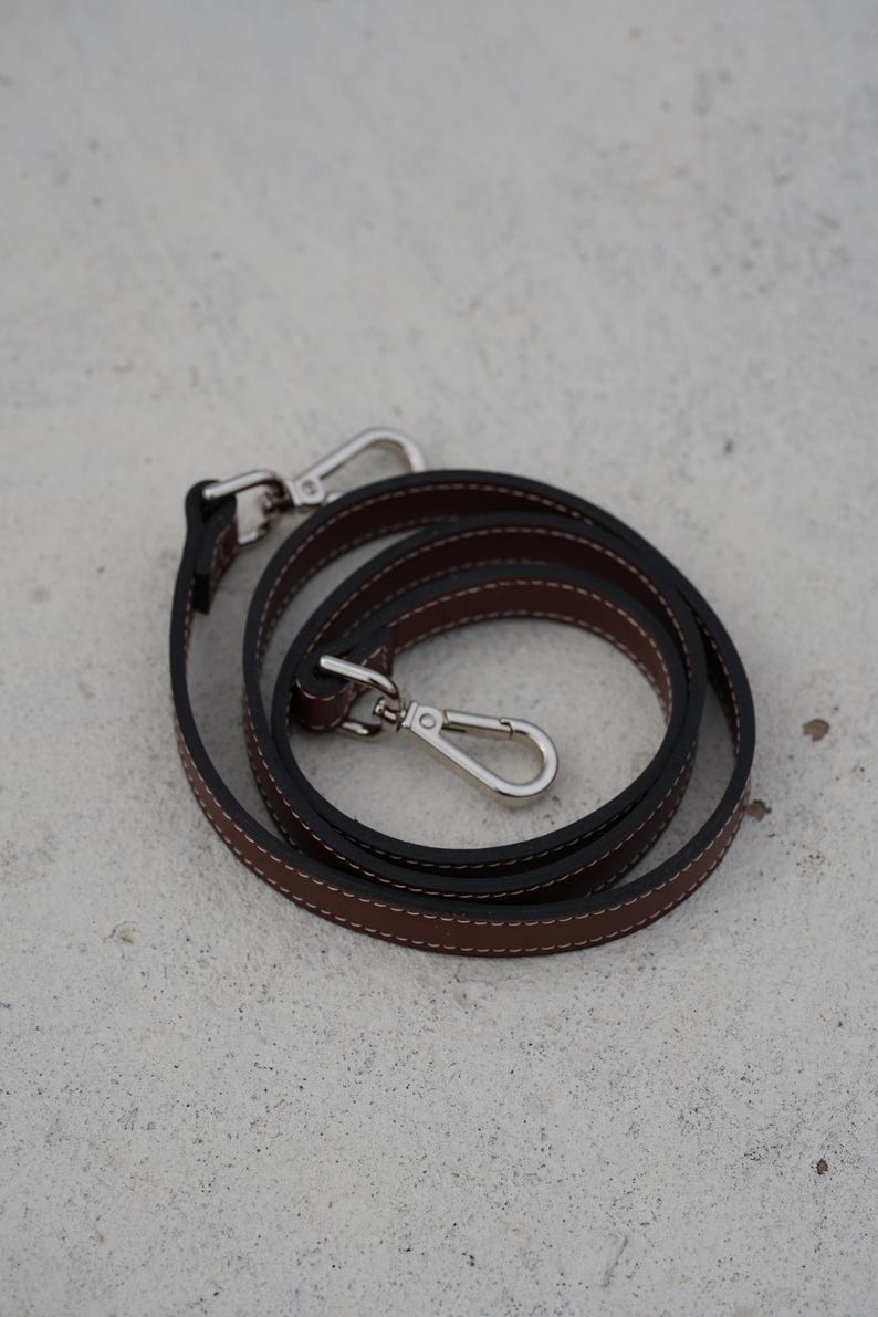 May include: A coiled brown leather strap with white stitching and black edges. The strap has two silver-colored metal clasps. The strap is designed for a bag or purse.