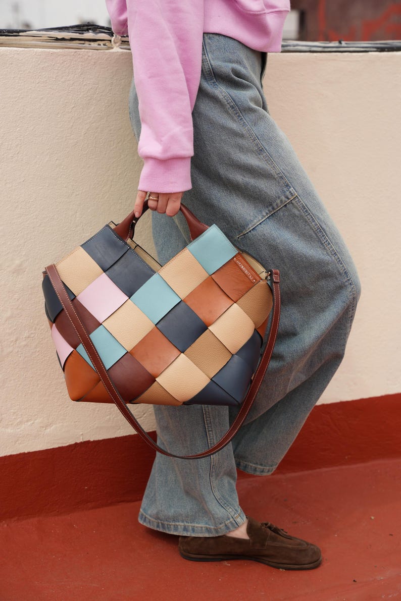 May include: A multi-colored woven leather tote bag with a brown shoulder strap. The bag features a patchwork design with squares of blue, tan, pink, and brown leather. The bag is held by a person wearing a pink sweatshirt and blue jeans.