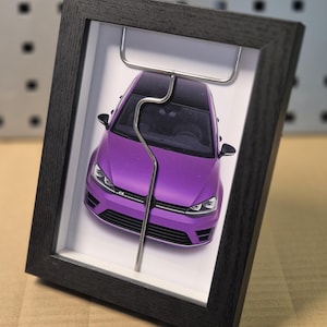 Peut inclure: Une voiture miniature violette avec un toit noir, encadrée dans un cadre en bois noir. La voiture est maintenue par un fil métallique argenté. Le fond est blanc.