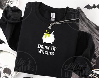 Op de afbeelding: Een zwart sweatshirt met lange mouwen en een witte afbeelding van een ketel waar limoengroene vloeistof over de rand stroomt. Onder de ketel staat de tekst "DRINK UP WITCHES" in een wit, gestileerd lettertype. Een zilveren ketting ligt op de stof.