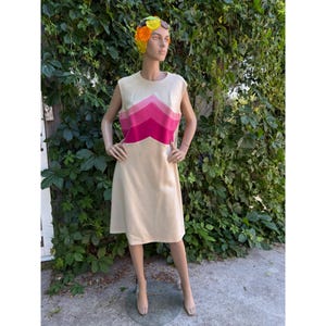 May include: A sleeveless cream-colored dress with a chevron design in shades of pink. The dress is knee-length and displayed on a mannequin. The mannequin is wearing a floral headpiece. The background features green foliage.