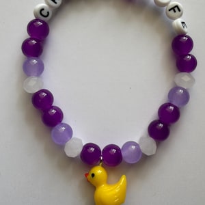 Beaded bracelet featuring purple, white, and translucent beads with white letter beads spelling CRUISE LIFE and a yellow rubber duck charm.