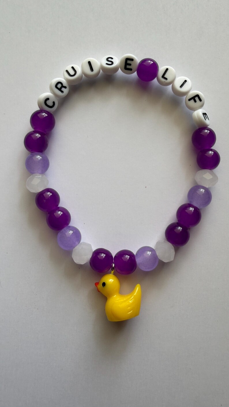 Beaded bracelet featuring purple, white, and translucent beads with white letter beads spelling CRUISE LIFE and a yellow rubber duck charm.