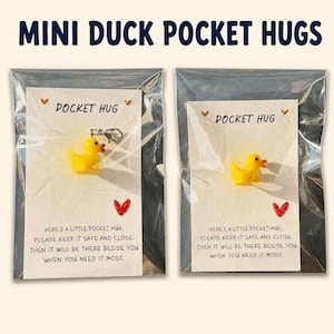 May include: Two mini yellow duck charms packaged in clear plastic sleeves on white cards. Each card features the text POCKET HUG, a red heart, and a poem: HERE'S A LITTLE POCKET HUG, PLEASE KEEP IT SAFE AND CLOSE. THEN IT WILL BE THERE BESIDE YOU WHEN YOU NEED IT MOST.